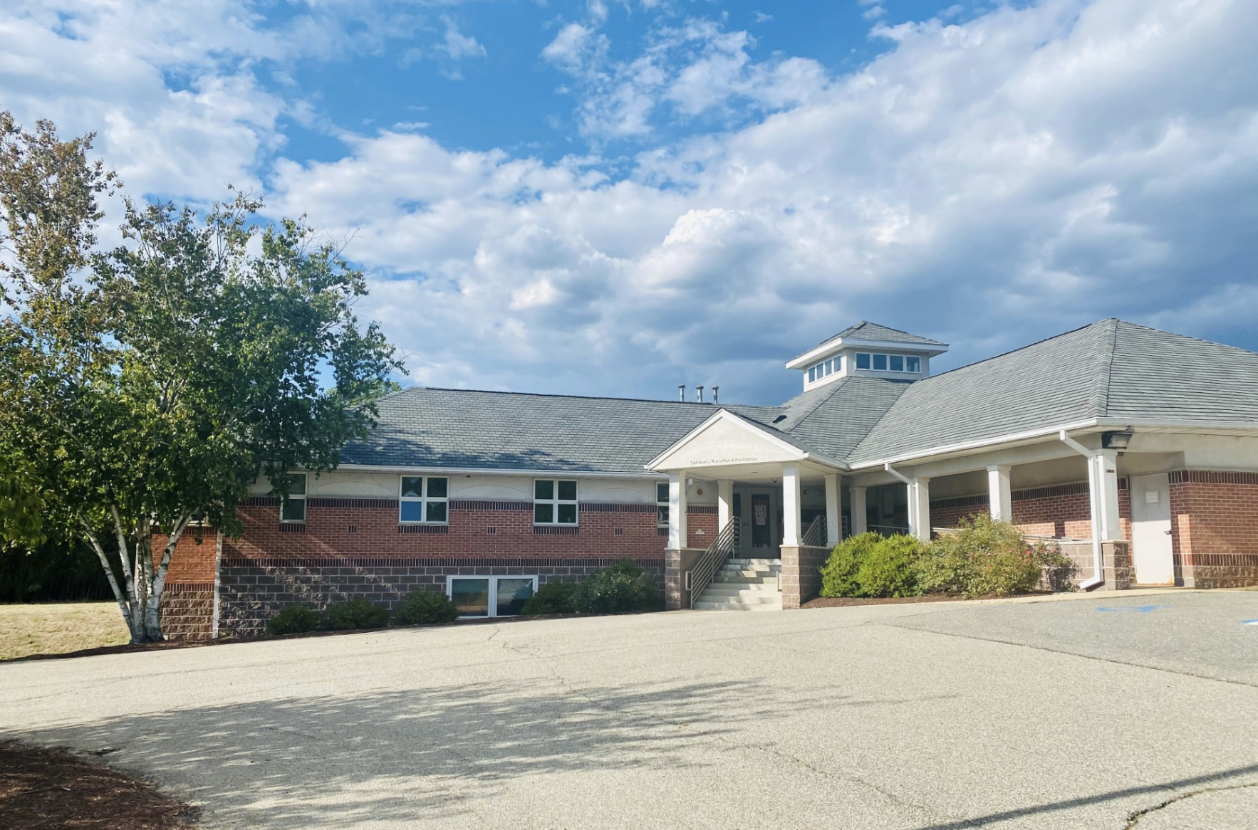 Front view of a one-story brick behavioral health facility with a covered entrance, white columns, and surrounding trees under a blue sky with scattered clouds.